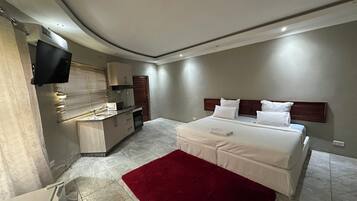 A modern bedroom with a large bed, a red rug, a built-in kitchen with a microwave, and a TV mounted on the wall.