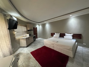 A modern bedroom with a large bed, a red rug, a built-in kitchen with a microwave, and a TV mounted on the wall.