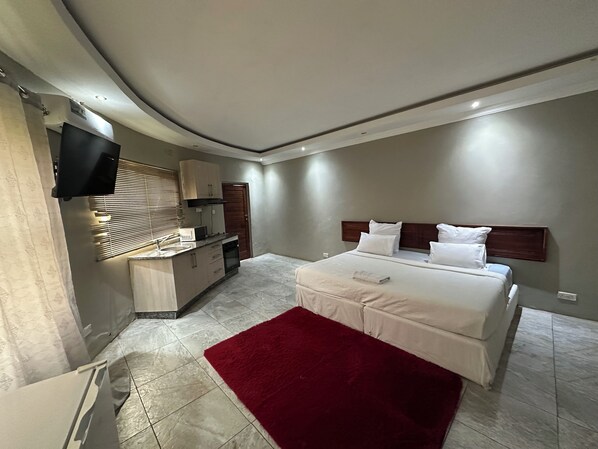A modern bedroom with a large bed, a red rug, a built-in kitchen with a microwave, and a TV mounted on the wall.