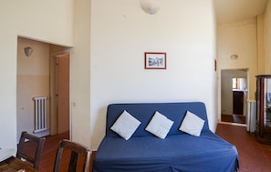 Ein Zimmer mit einem blauen Schlafsofa, einem Heizkörper, einem gerahmten Bild an der Wand und einer Tür zu einem anderen Raum.