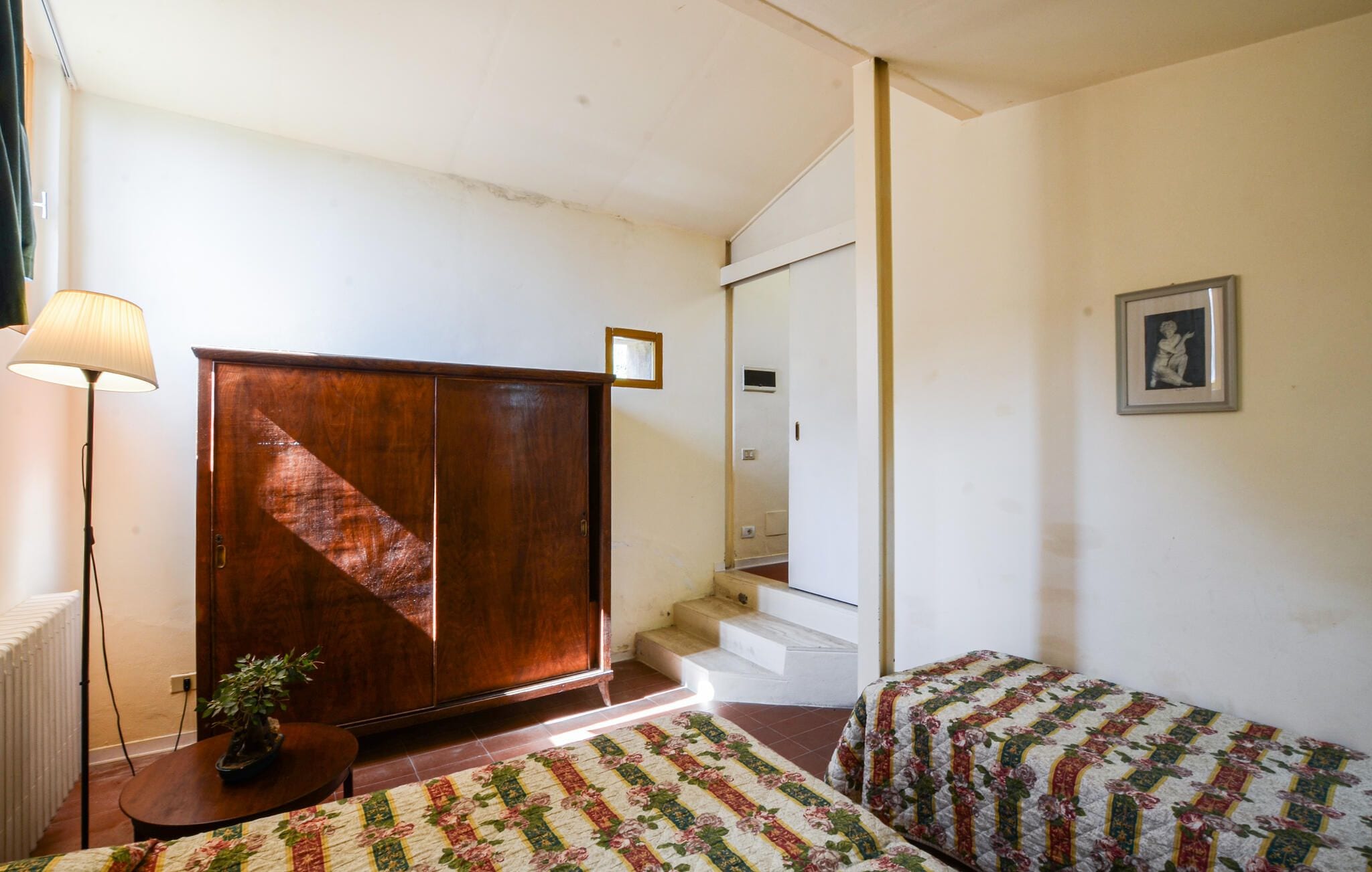 A room with a wooden wardrobe, a small table with a plant, a patterned bedspread, and a framed picture on the wall.