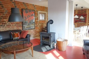 Ein Wohnzimmer mit Backsteinwand, einer schwarzen Couch, einem Glastisch und einem schwarzen Kamin. Im Hintergrund ist ein Essbereich zu sehen.