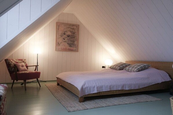 Une chambre avec un plafond en pente, un lit recouvert d’une couvre-lit de couleur claire, une chaise à motifs floraux et une œuvre d’art accrochée au mur.