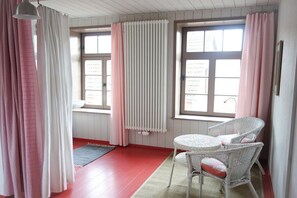 Une pièce avec un plancher en bois rouge, une table en osier blanche avec deux chaises, un radiateur et des rideaux roses.