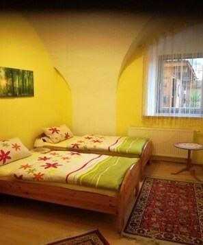 Una habitación con dos camas de madera, una mesita redonda pequeña y un ventanal con cortinas.