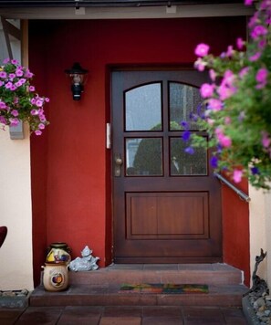 Eine rote Tür mit Glasfenster, ein kleiner Podeststufenaufgang davor und ein hängender Blumentopf mit violetten Blüten.