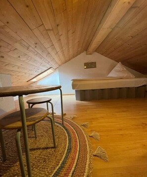 Ein kleines, gemütliches Schlafzimmer unter dem Dach mit Holzbalkendecke, einem Bett, einem Tisch mit Stuhl und einem bunten Wollteppich.