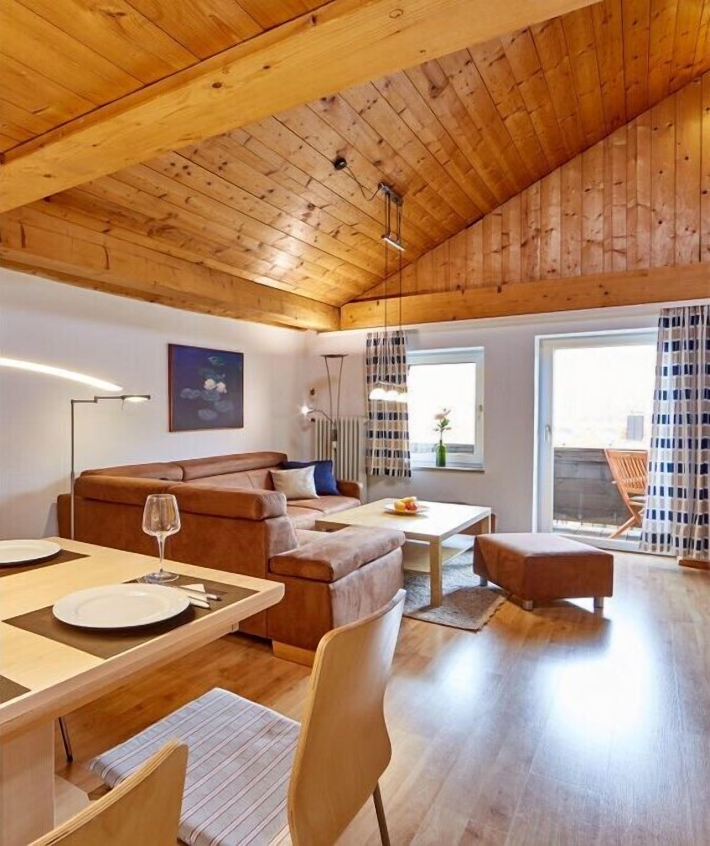 A spacious living area with a wooden ceiling, a sofa, a dining table with chairs, and a view through large windows.