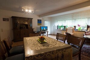 Een eetruimte met een tafel voor vier personen, omringd door rieten stoelen, een kast met glazen deuren en een televisietoestel op een standaard.