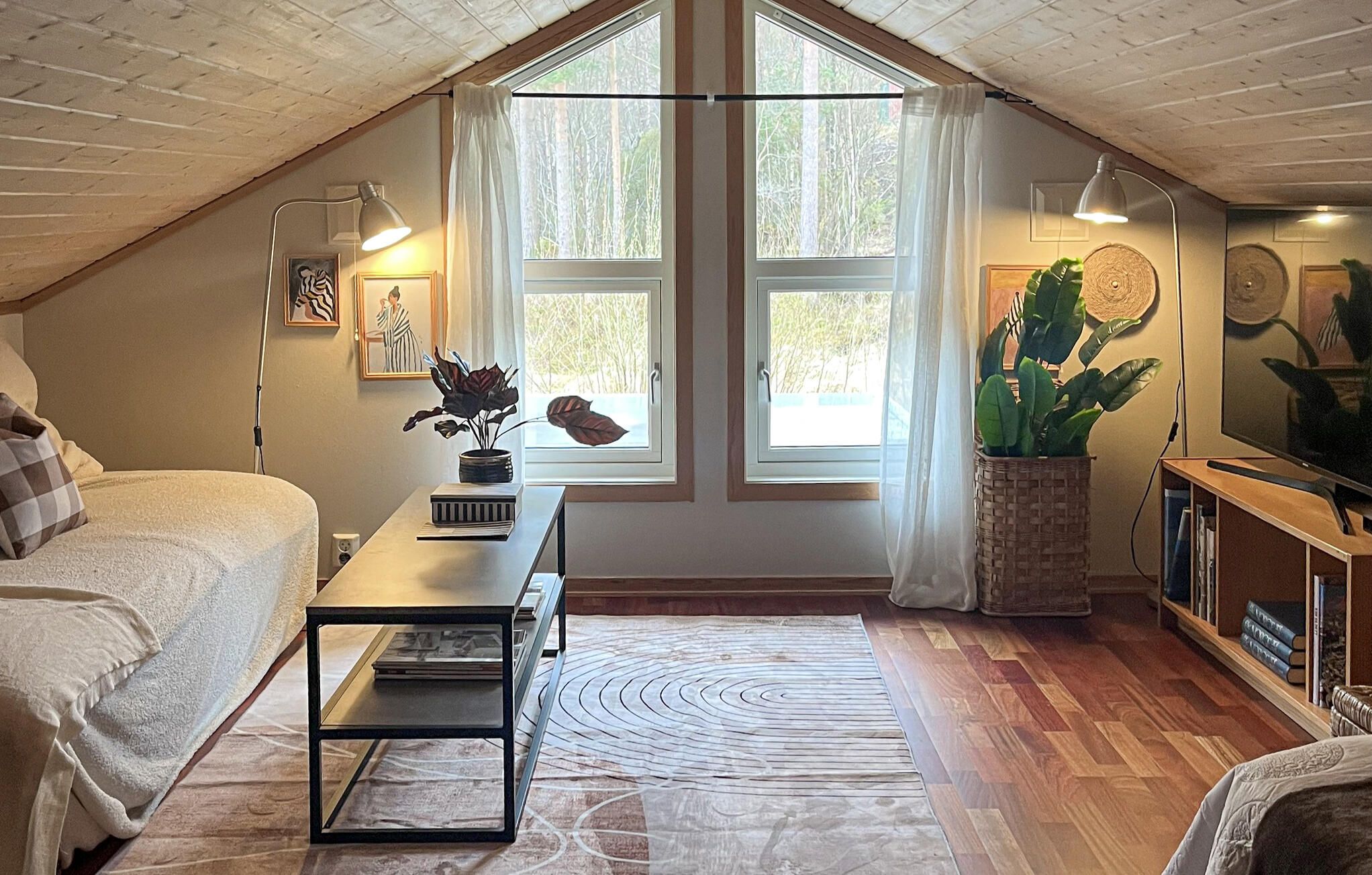 A cozy loft bedroom with a sloped ceiling, a large window with curtains, a bed with a checkered pillow, a side table with a lamp, a potted plant, and a television.
