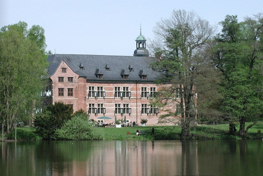 A large building with a prominent tower, situated next to a calm lake with trees around.
