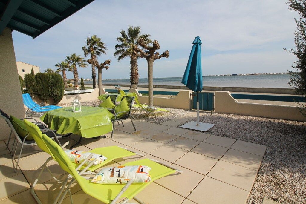 An outdoor seating area with green lounge chairs and a table, overlooking a body of water with palm trees and a clear sky.