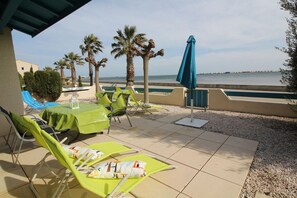 An outdoor seating area with green lounge chairs and a table, overlooking a body of water with palm trees and a clear sky. - Holiday home in Gruissan with swimming pool (Gruissan)