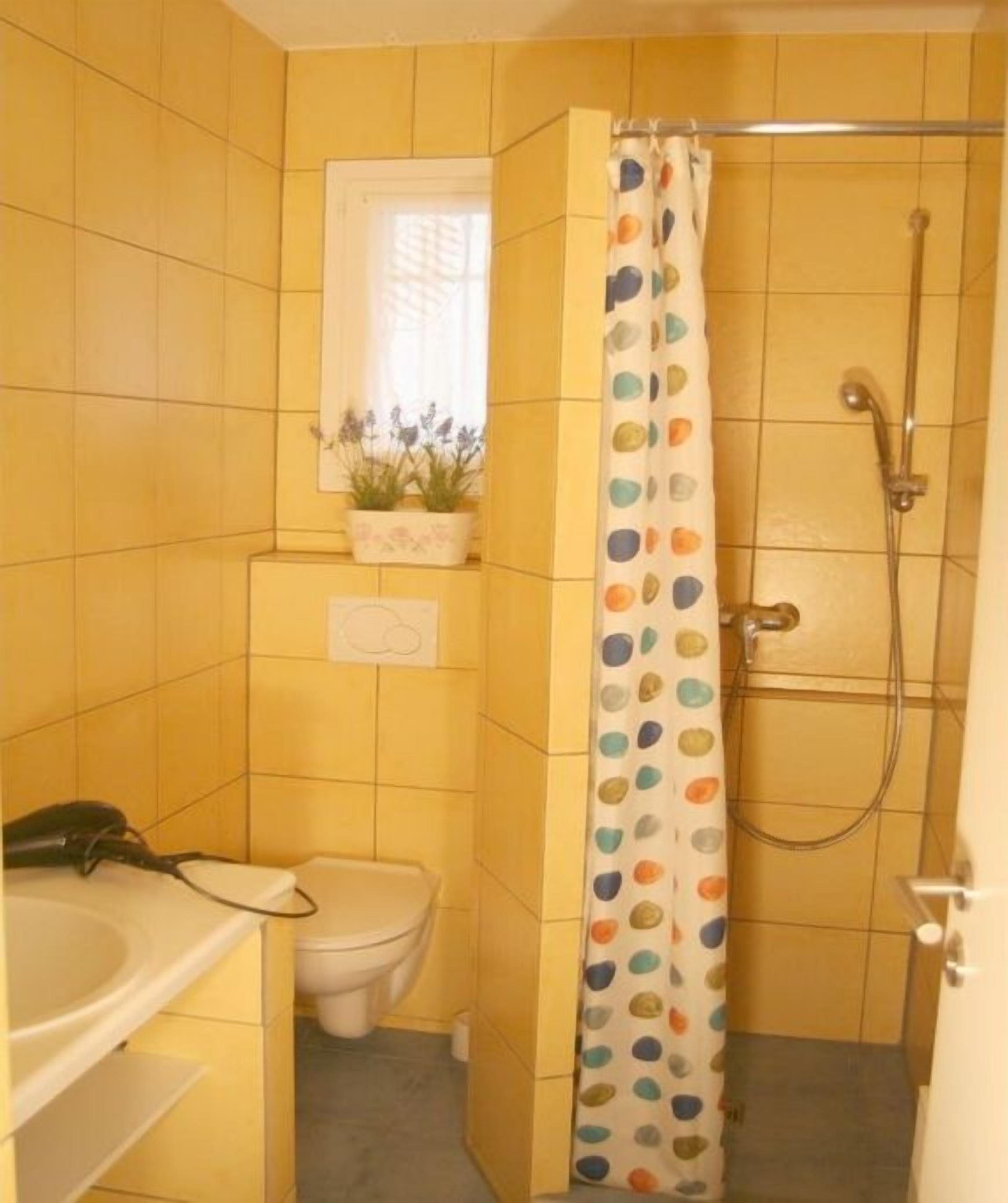 Une salle de bain avec des carreaux jaunes, des toilettes, un lavabo, une douche avec un rideau à pois et une fenêtre avec une plante.