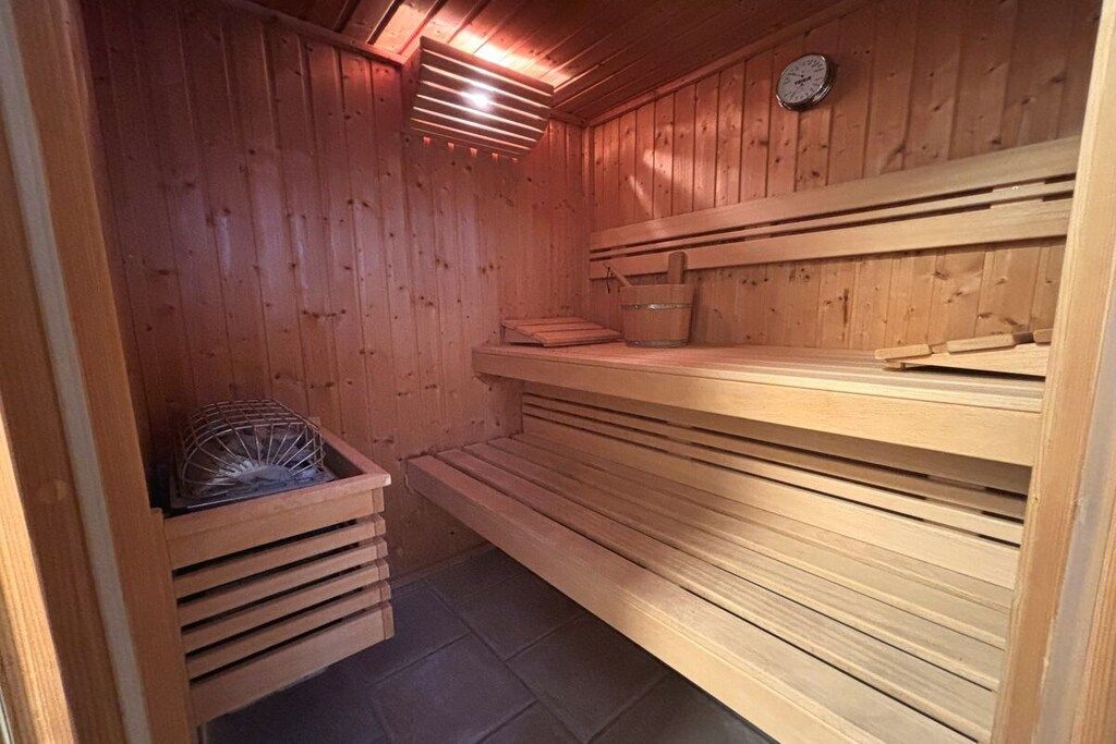 A sauna with wooden walls and benches, a clock, and a small shelf with a bucket.