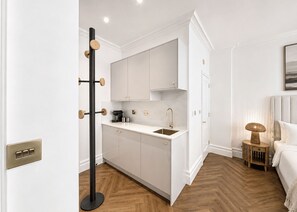 A compact hotel room with a kitchenette, a bed, a bedside table with a lamp, and a view of the outdoors. - Nestoya House (London)