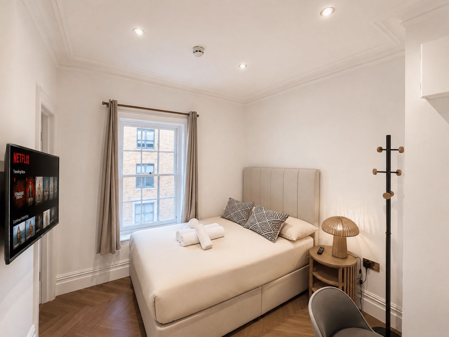 Quarto de hotel com cama, televisão na parede, janela com cortinas, luminária de cabeceira e um cabideiro.