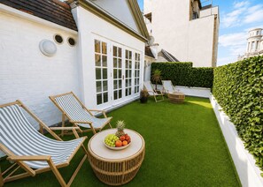 A patio with two striped lounge chairs, a round wicker table with a fruit bowl, and a hedge. - Nestoya House (London)
