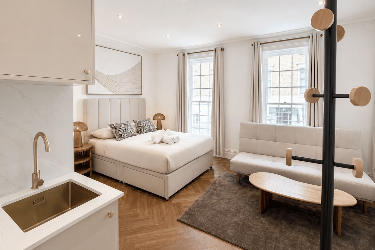A modern hotel room with a bed, a sofa, a small table, a bathroom with a sink, and large windows.