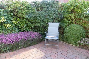 Ein Stuhl in einem Gartenbereich mit einem gepflasterten Weg, umgeben von dichtem Grün und einem kleinen Busch mit violetten Blüten.