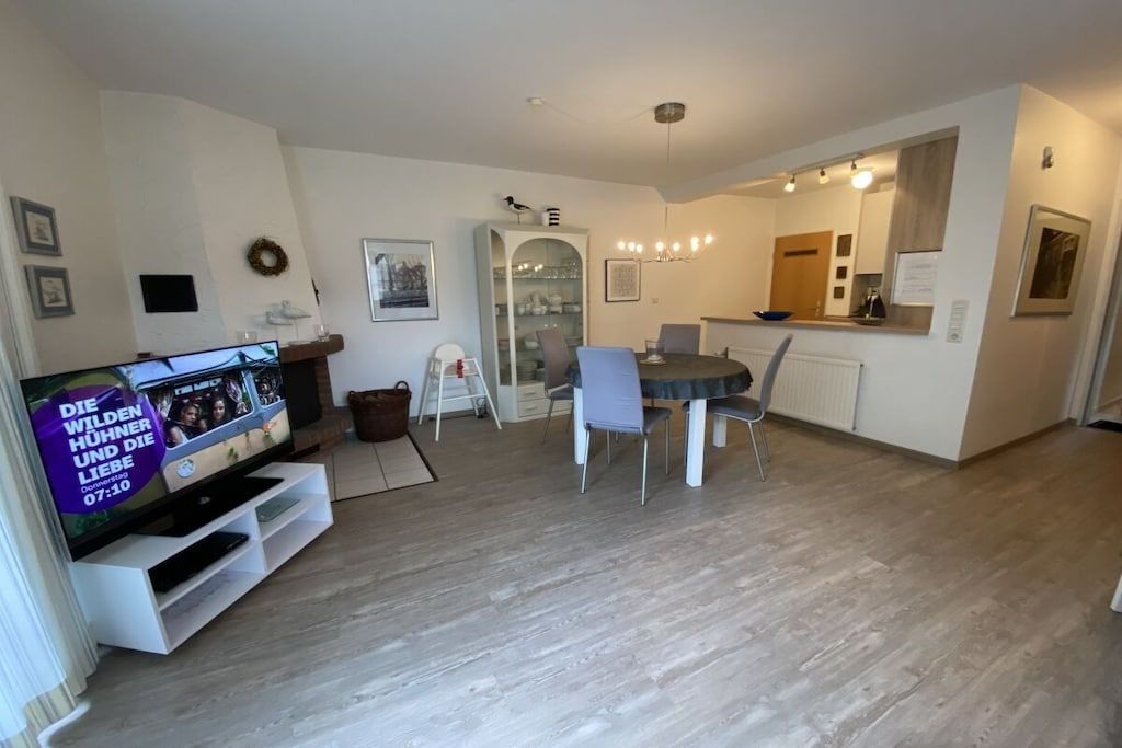 Una sala moderna con un televisor de pantalla plana, un comedor con una mesa redonda y sillas, y una cocina con refrigerador y encimera.