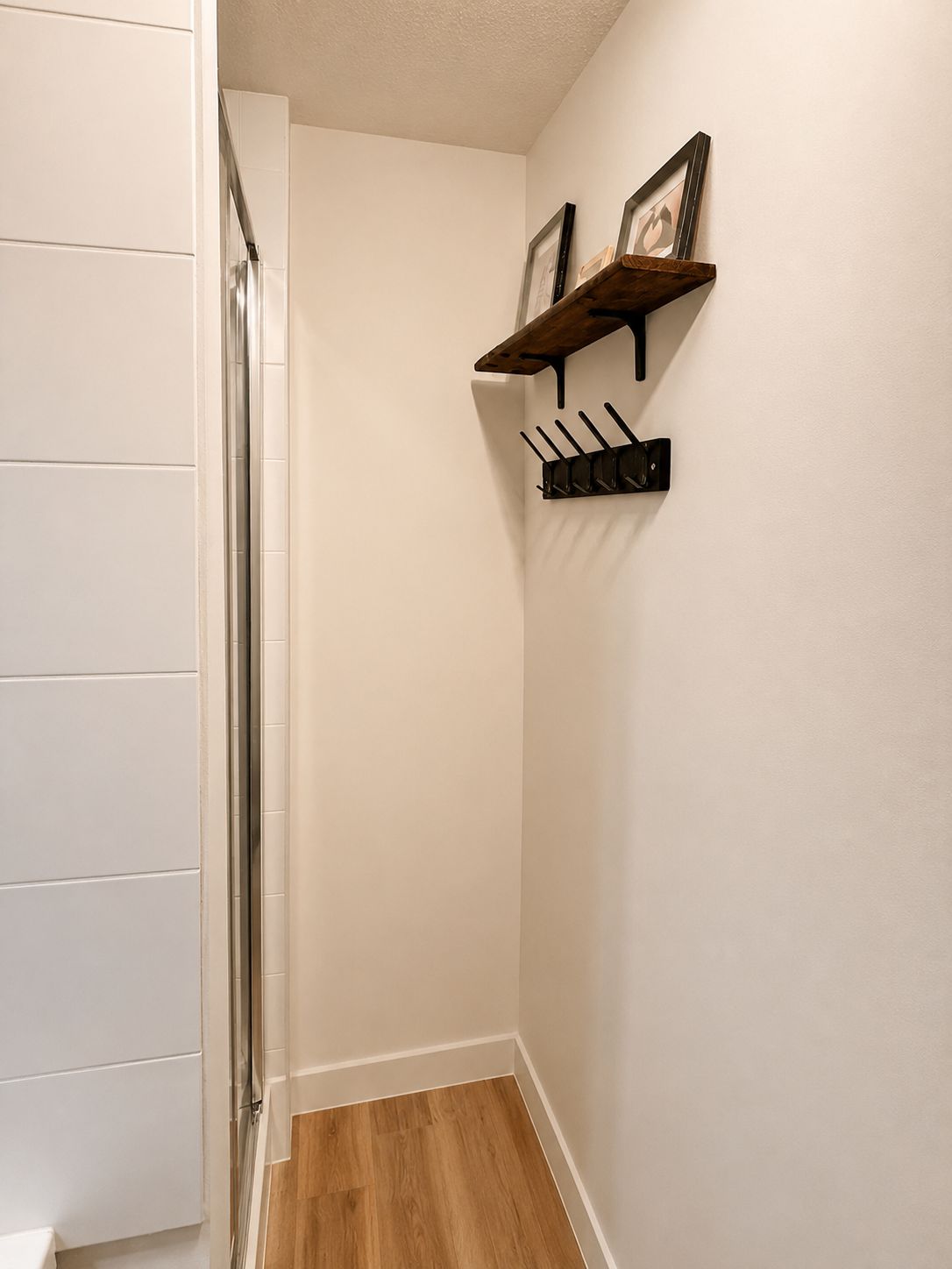 Un couloir étroit doté d’un plancher en bois, de murs blancs et d’une étagère noire fixée au mur, munie de crochets.