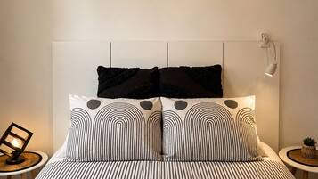 Un dormitorio con cama, almohadas, una mesita de noche, una lámpara, un estante con objetos decorativos y una lámpara de pared.