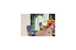 Une terrasse en bois avec deux chaises et une table, une fenêtre donnant sur un beau paysage et un parterre de fleurs roses.