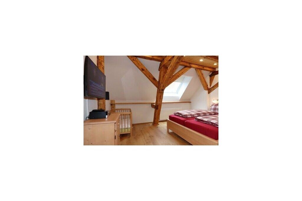 Un dormitorio con techo inclinado, vigas de madera, una cama con colcha roja, una cuna de madera pequeña y un televisor sobre un mueble.