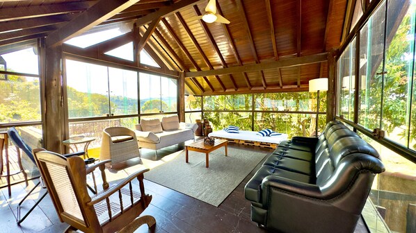 Una habitación amplia con techo de madera, ventanas grandes con vistas al exterior, un sofá, una silla y una mesita.