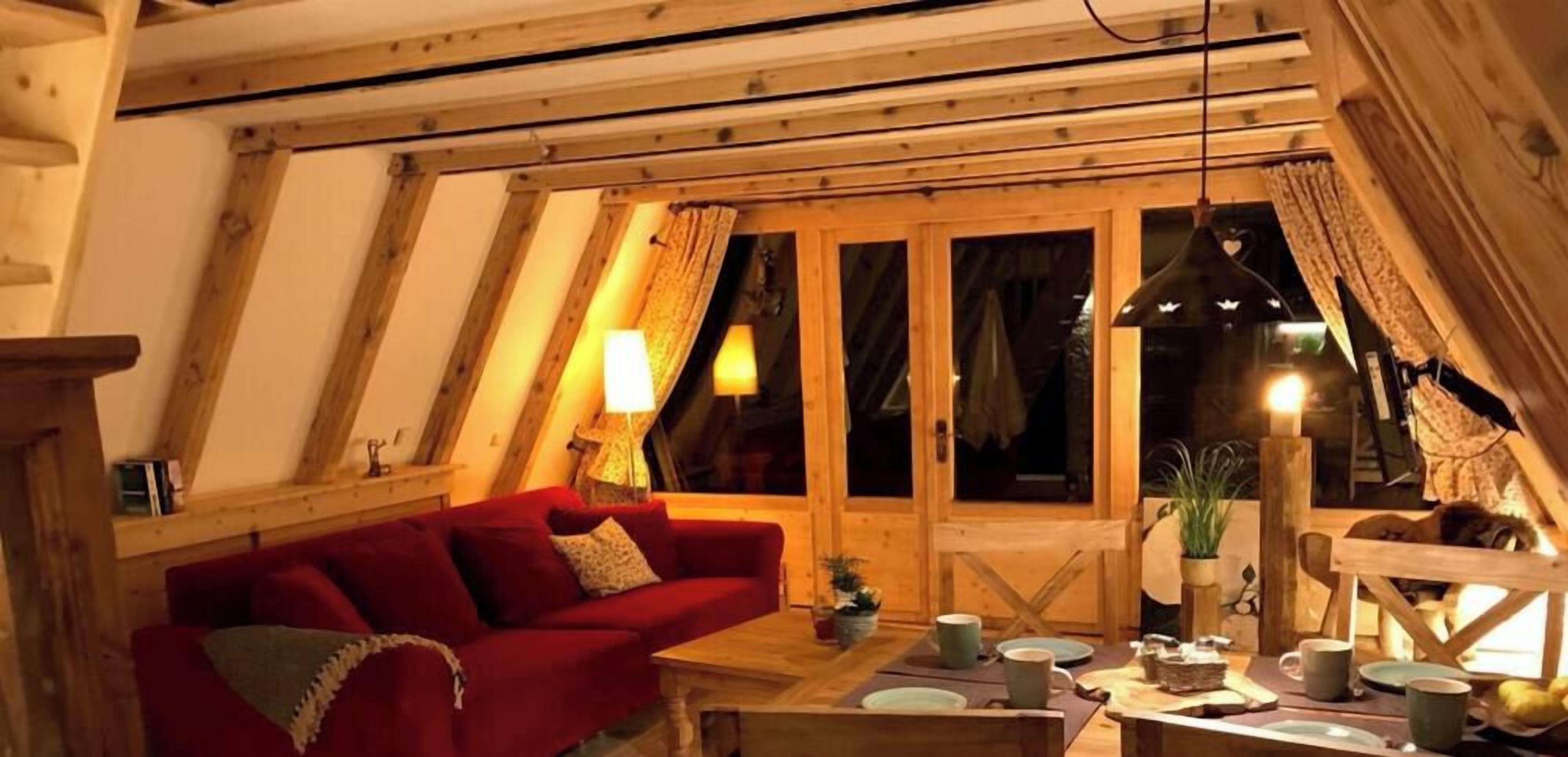 A cozy room with a red sofa, wooden beams, a dining table with plates and cups, and a window with curtains.