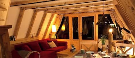 Una habitación acogedora con un sofá rojo, vigas de madera, una mesa de comedor con platos y tazas, y una ventana con cortinas.
