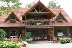 A two-story wooden building with a tiled roof, a balcony with flowers, and a front porch with chairs and tables. - Holiday apartment with 4 bedrooms (Pegnitz)