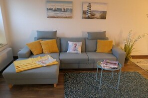 Un salon avec un canapé gris, des coussins jaunes, une petite table d’appoint avec des magazines et un tapis noir et blanc.