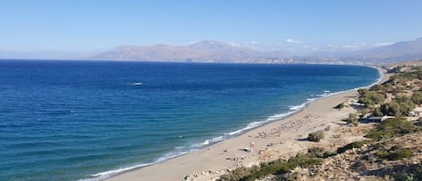 Un paisaje costero con playa, cielo despejado y montañas a lo lejos.