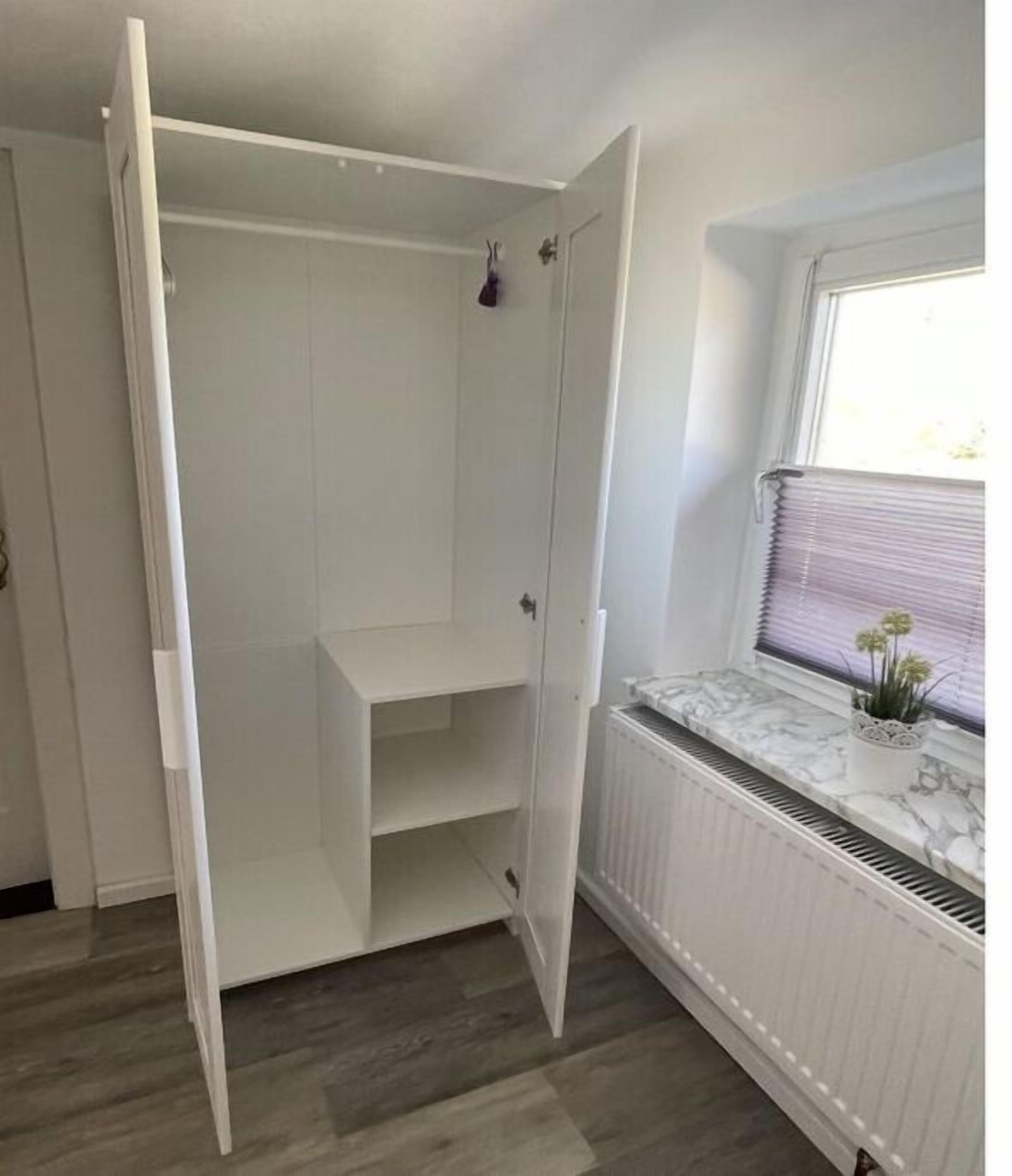 Une pièce avec un radiateur blanc, une fenêtre avec des stores et une armoire blanche dont les portes sont ouvertes.
