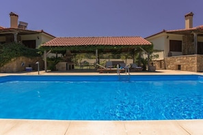 A swimming pool with a covered patio area, stone walls, and a clear blue sky.