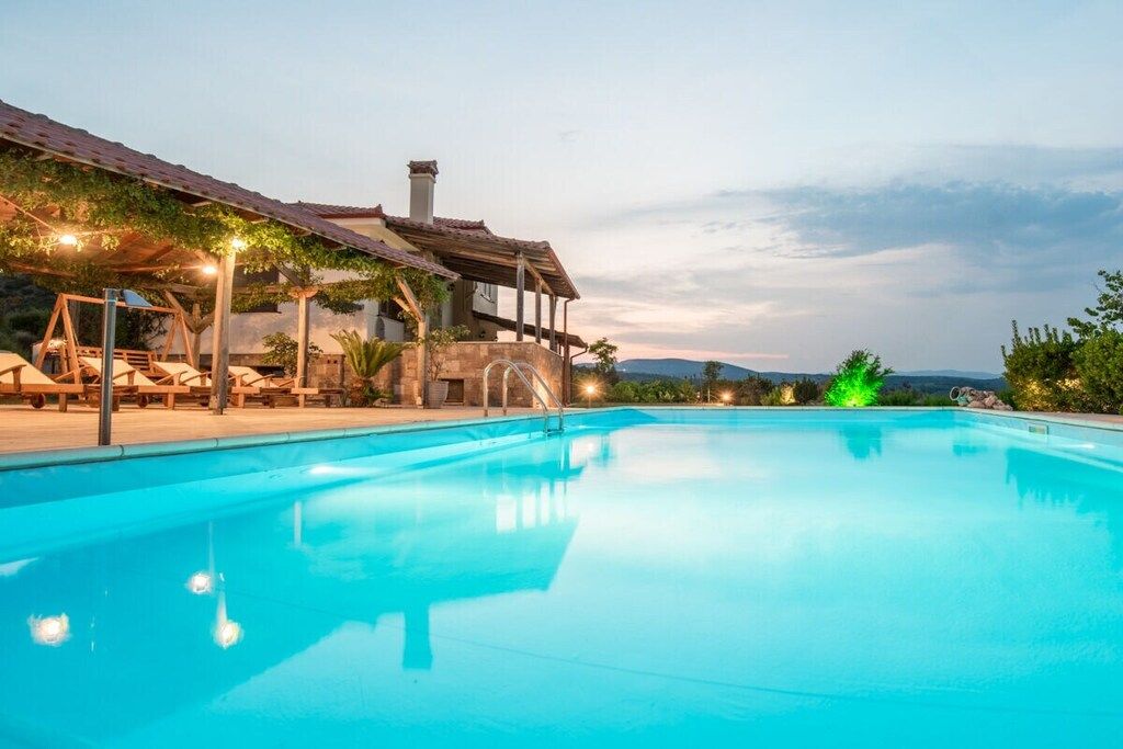 Una piscina con sillas de descanso y una zona cubierta, rodeada de vegetación y vistas a colinas distantes.