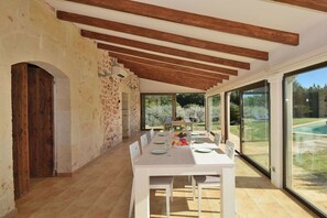 An open-plan dining area with a long table set for a meal, wooden beams on the ceiling, and large windows offering a view of the outdoors.