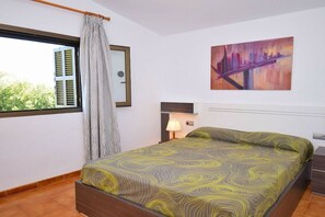 A bedroom with a bed, a window with shutters, a painting on the wall, and a nightstand with a lamp.