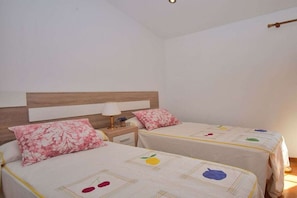 A bedroom with two beds, each with a floral pillow and a bedspread with fruit patterns. There is a nightstand with a lamp and a small table with a vase.