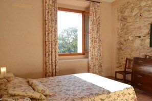 Ein Schlafzimmer mit einem Bett, einer Kommode, einem Fenster mit Vorhängen und einer Steinwand.