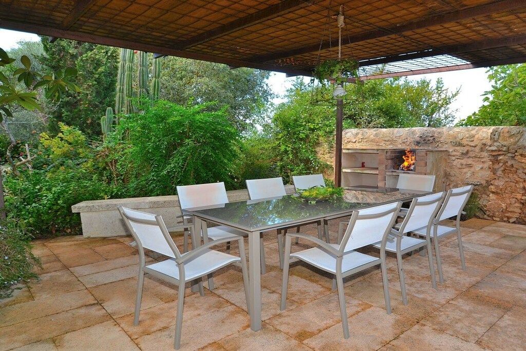 An outdoor patio with a stone table and chairs, a stone oven, and surrounding greenery.