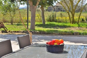 A table with a wicker basket containing fruit, surrounded by wicker chairs, set outdoors with trees and a clear sky in the background.
