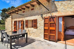 A stone building with a patio, a wooden table with chairs, and a bed visible inside.