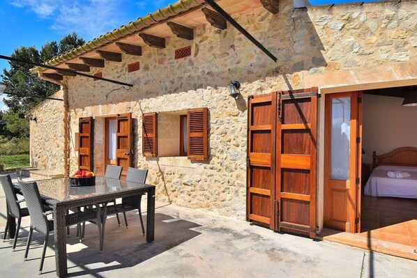 A stone building with a patio, a wooden table with chairs, and a bed visible inside.