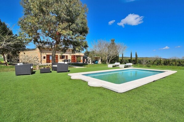 A pool with a surrounding grassy area, a building with a stone facade, and a large tree.