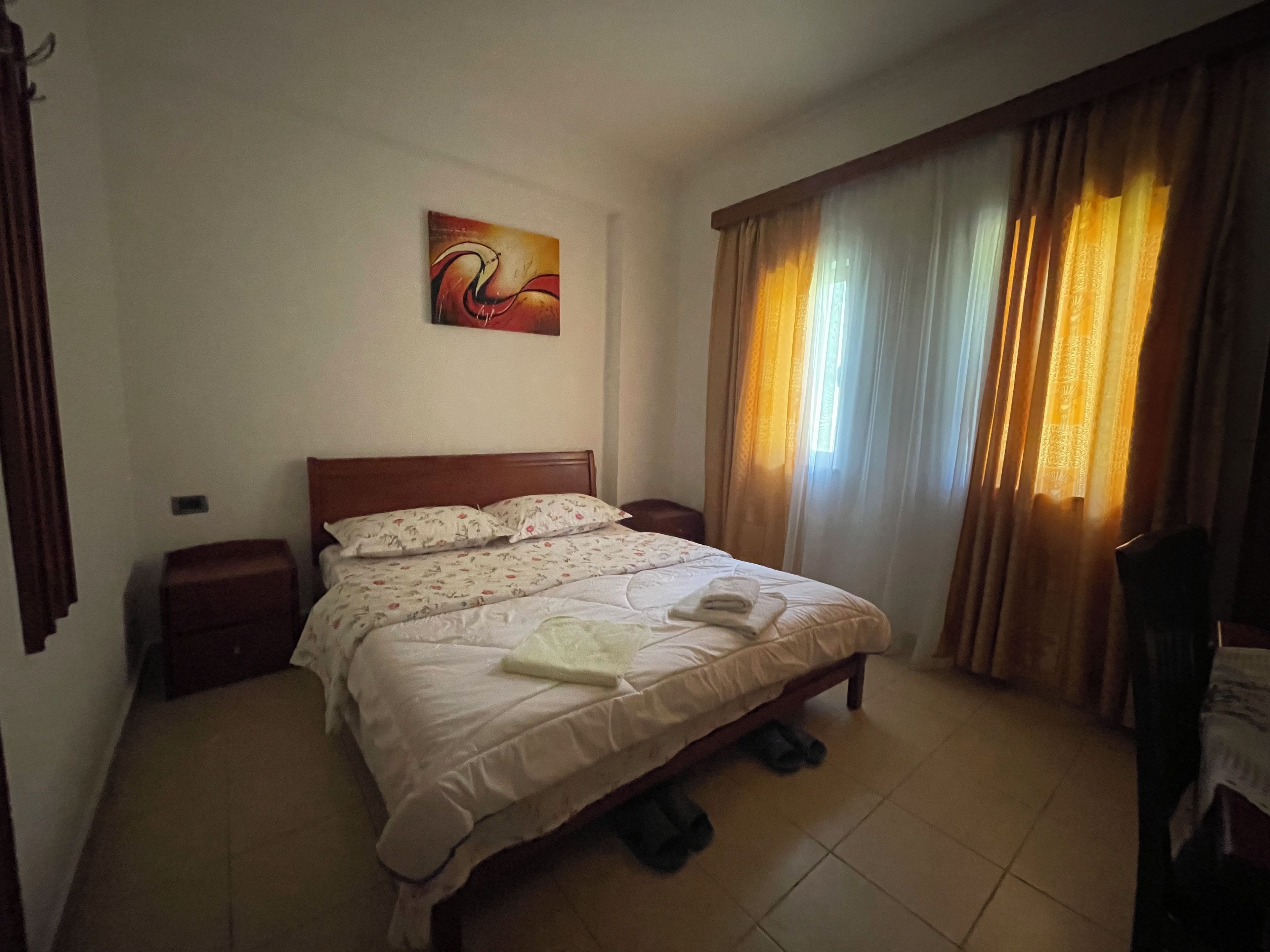 A hotel room with a bed, a nightstand, a wall-mounted painting, and a window with curtains.