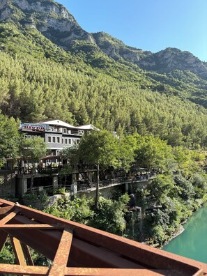 Un restaurante junto al río con terraza al aire libre que ofrece vistas a un valle arbolado y montañas al fondo.