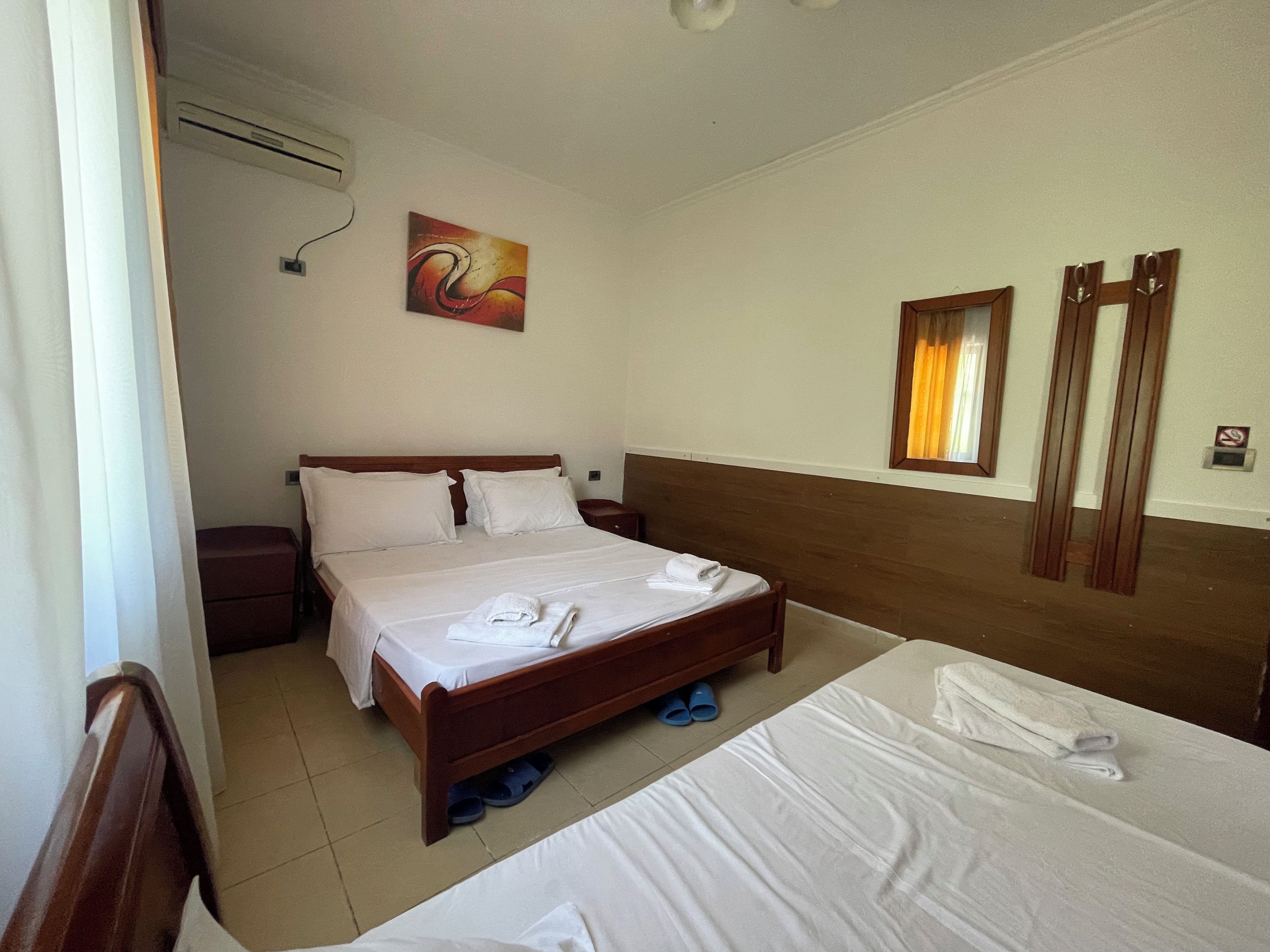 Habitación de hotel con cama doble, cama individual, mesita de noche, aire acondicionado instalado en la pared, un cuadro y una ventana con cortinas.